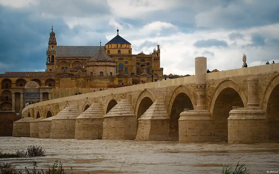Die Römische Brücke von Córdoba über den Guadalquivir, dahinter die Mezquita-Catedral mit ihrem Turm unter bewölktem Himmel